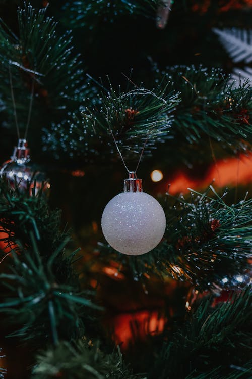 Closeup of white decorative Christmas ball hanging on branches of artificial fir tree on blurred background
