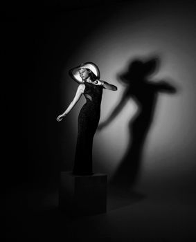 Black and white of slim woman model in stylish long dress and hat standing with raised arms in dark studio against shadow on wall