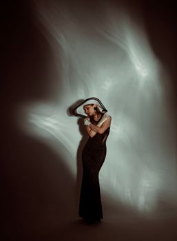 Full length of charming female in stylish black dress and hat standing with crossed arms in dark studio and illuminated by light