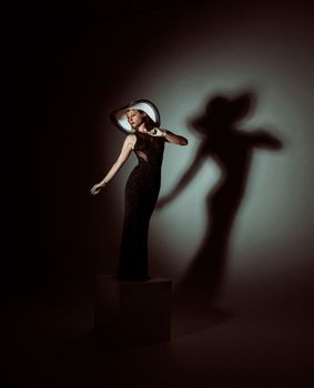 Full body of stylish woman in long black dress and hat standing in dark studio and looking away