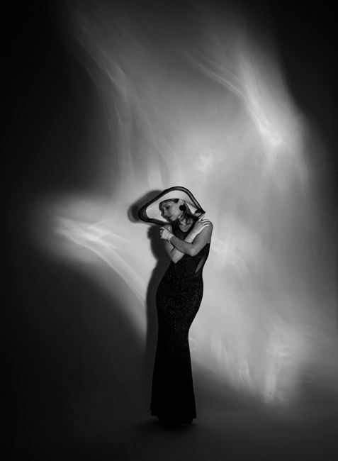 Black and white of model in long stylish dress and hat standing with crossed arms in dark studio and illuminated by glowing light