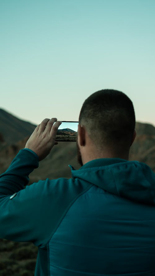 Back View of a Man in a Blue Jacket Taking a Photo of a Mountain