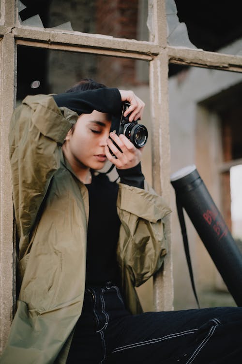 Concentrated young female photographer in casual outfit taking pictures on professional photo camera while standing near broken window of abandoned building in daylight