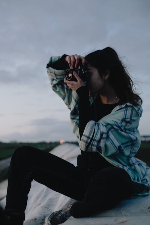 Free Concentrated young female photographer in casual clothes taking pictures on modern photo camera while sitting on roof in countryside at peaceful twilight Stock Photo