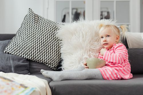 Free Charming toddler in striped dress sitting comfortably on a sofa, holding a bowl, surrounded by pillows. Stock Photo