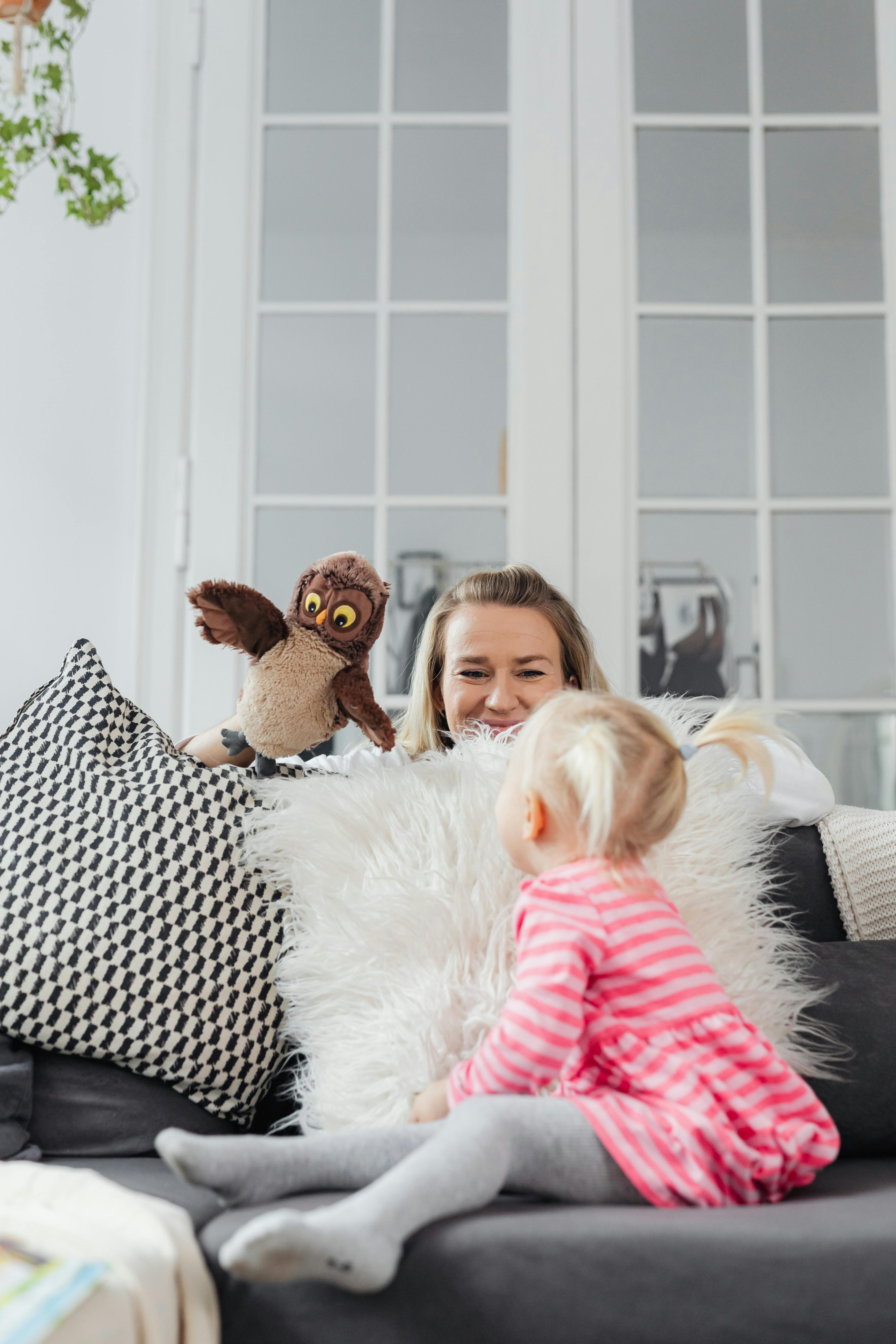 A mother uses an owl hand puppet to entertain her daughter in a cozy living room setting.