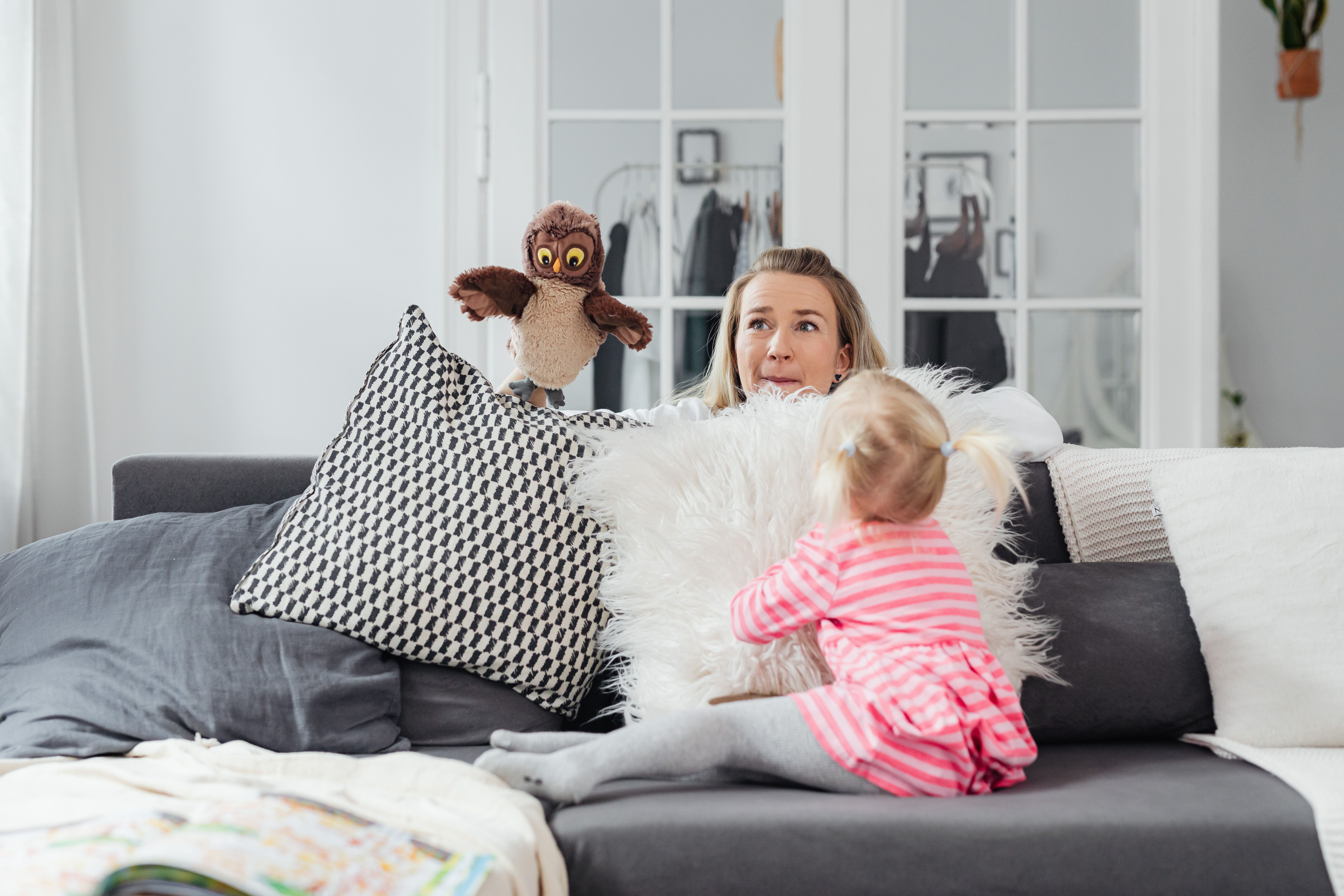 A woman and her daughter play with an owl puppet on a cozy couch in a bright living room.