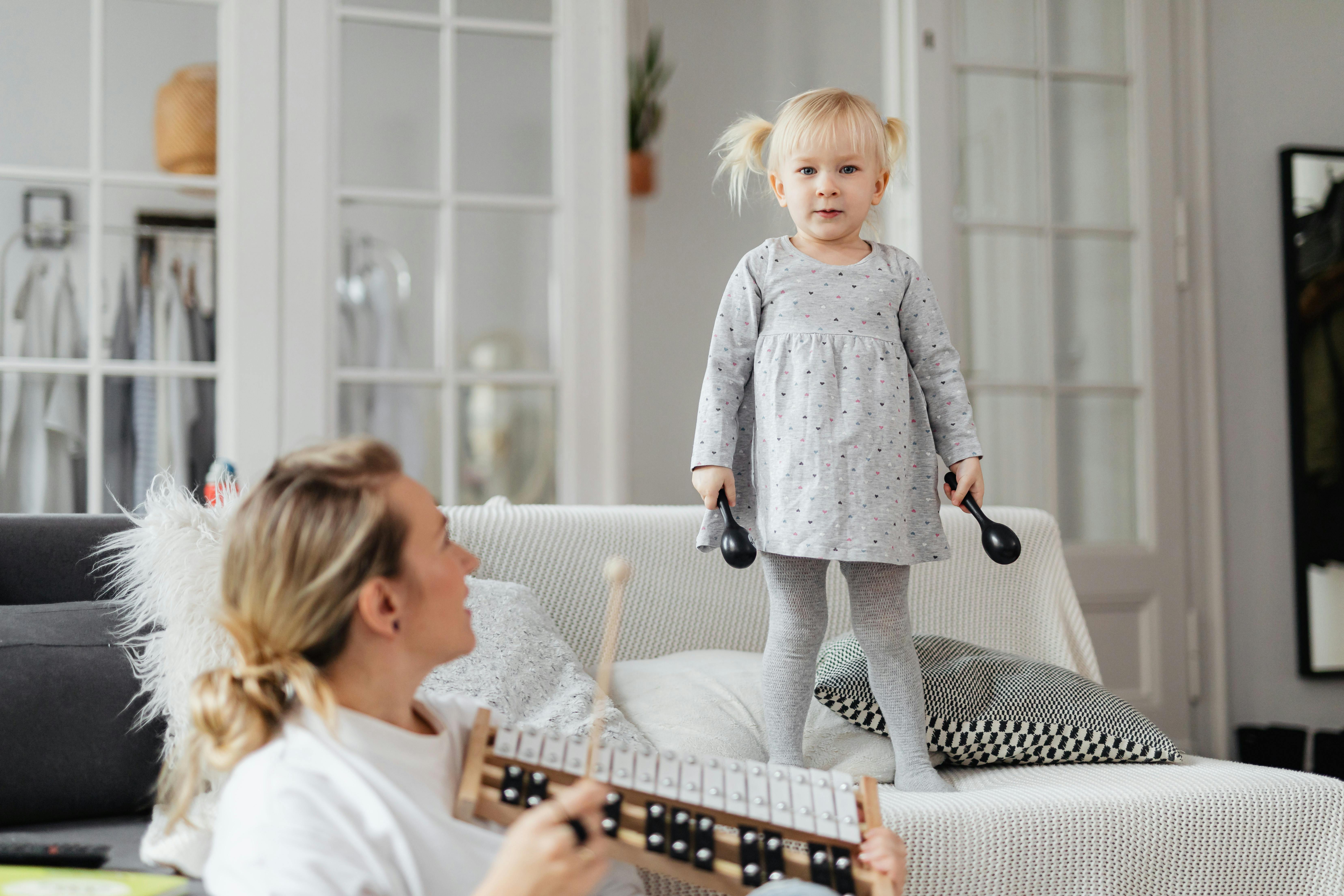 Young girl standing on couch holding percussion instruments, enjoying playful music time with adult nearby. | BocaVibe