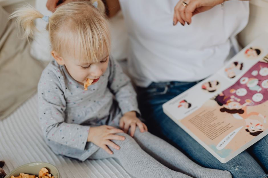 Cute toddler with ponytails enjoying a snack next to a book and parent. Cozy indoor scene.