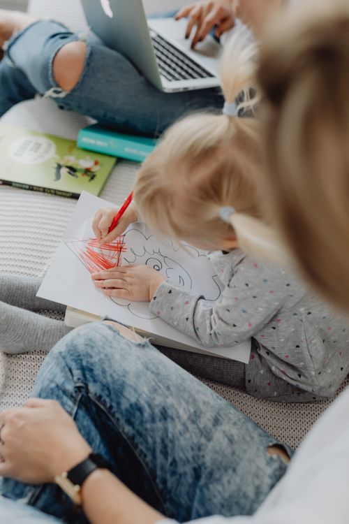Free Young child coloring with a red crayon while under adult supervision in a cozy home setting. Stock Photo
