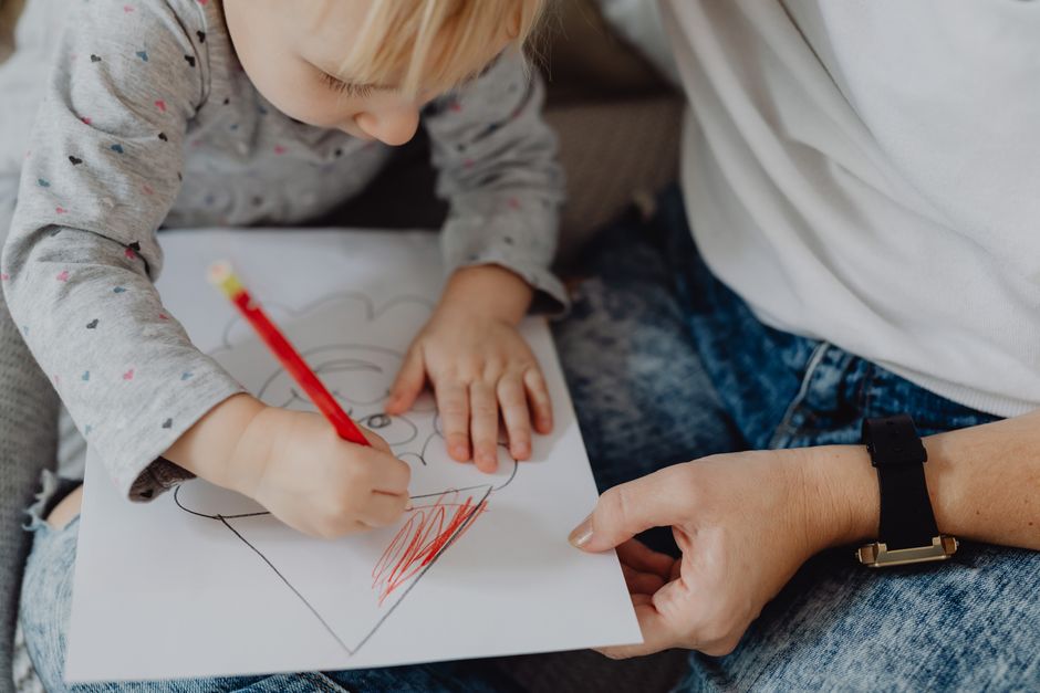Photo by https://kaboompics.com/ on Pexels A young child focused on coloring a drawing with a red pencil under adult supervision. - Photo by https://kaboompics.com/ on Pexels