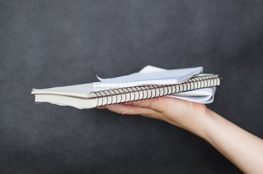 这张特写照片中，一名女子的手拿着一叠螺旋装订的笔记本和纸张，背景是暗色的