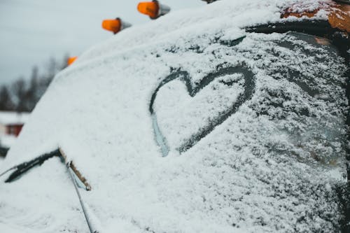 Heart symbol drawn on snowy windshield of truck with lamps parked on rural area with building on cold winter day