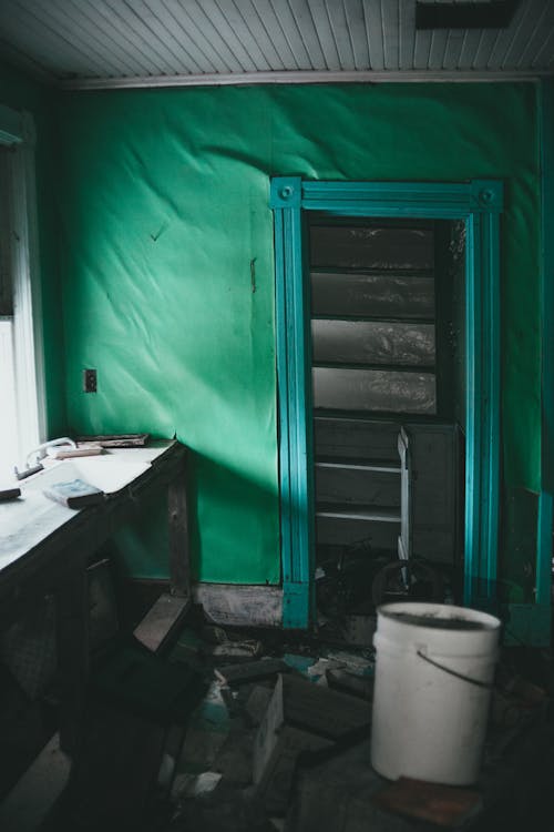 Interior of shabby room of rural cottage with plastic bucket om messy floor near weathered wooden table against green wall