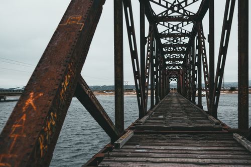 Free Aged bridge with wooden walkway and metal fences with rust over rippled ocean against mountains Stock Photo