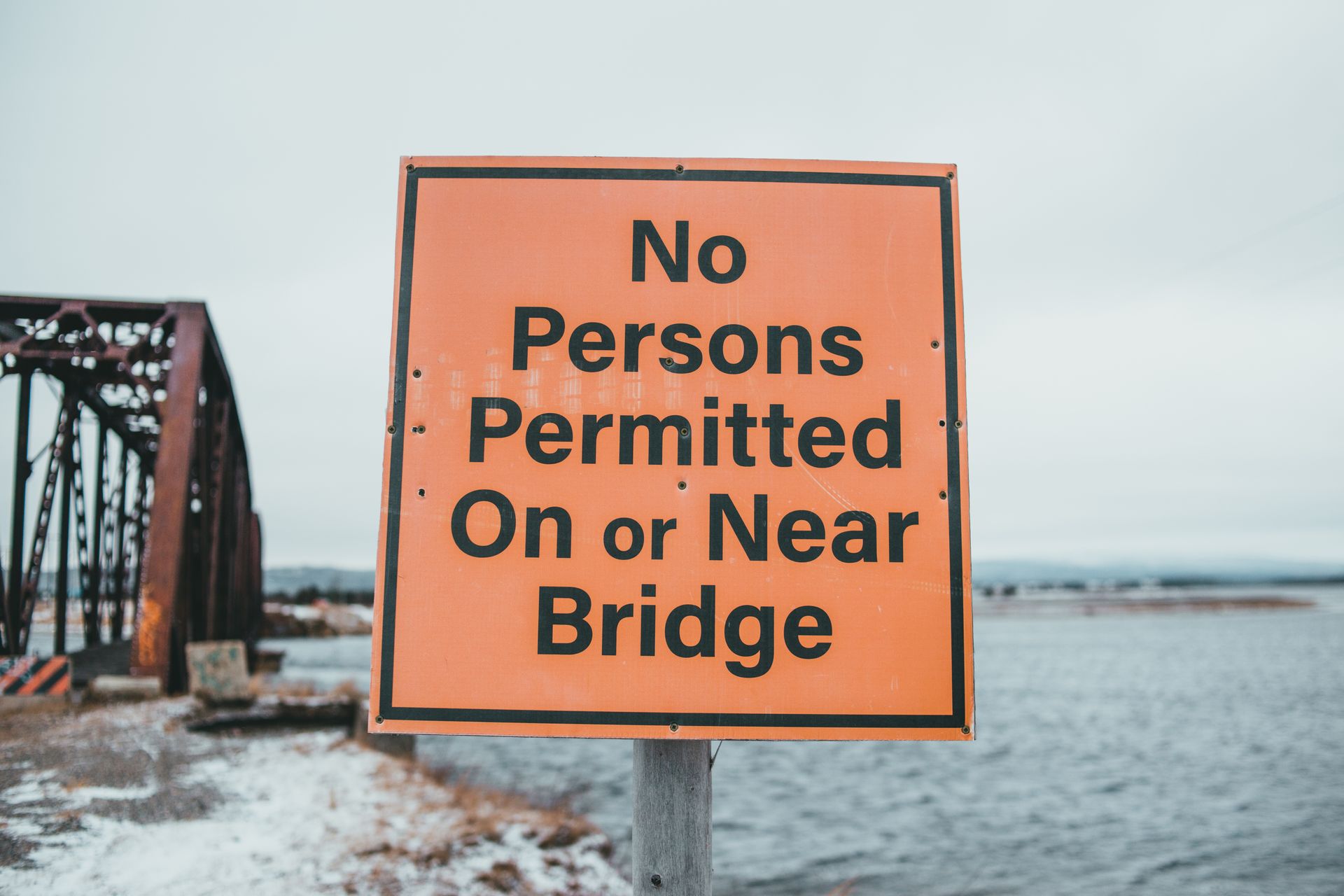 A bright orange sign warns against access near a bridge over a cold seacoast.