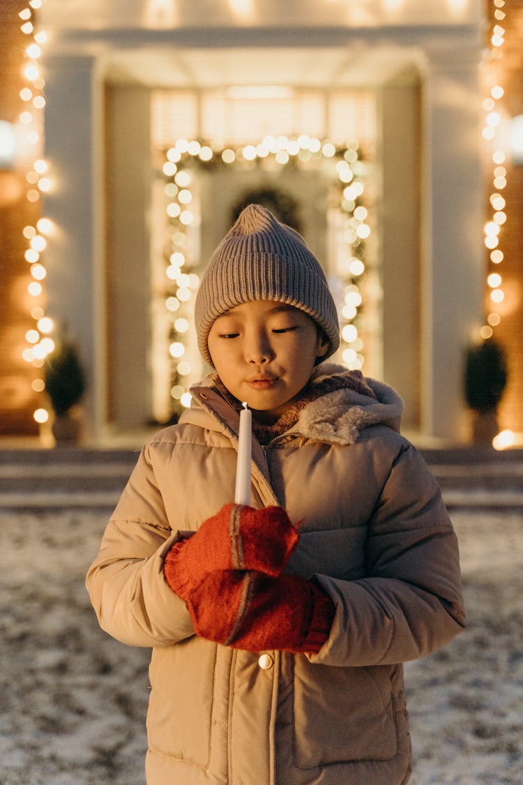 A Child In Winter Clothes Blowing Out A Candle