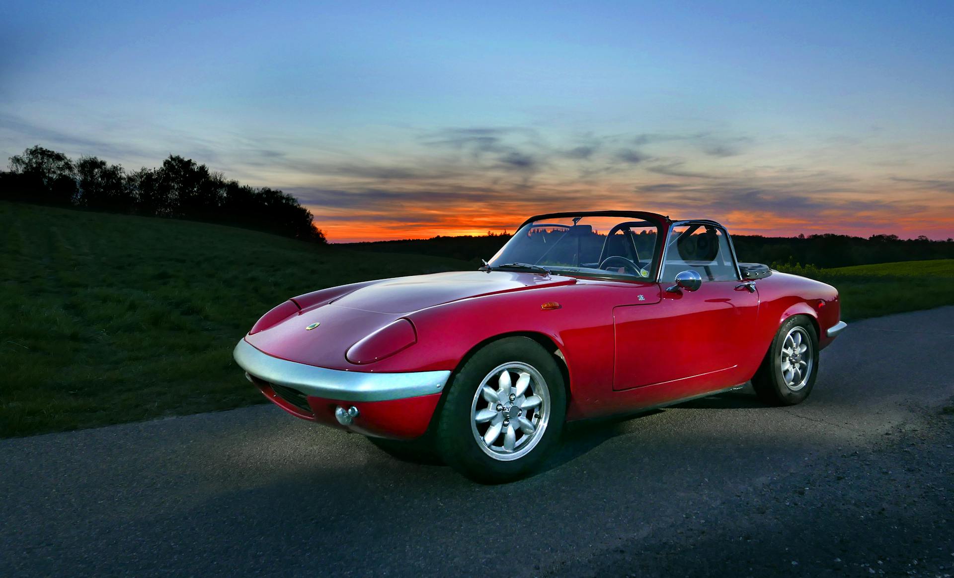 A classic red convertible sports car parked on a road against a stunning sunset backdrop.