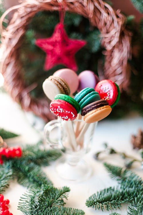 Sweet multicolored macaroons on sticks in cup placed on table near Christmas decorations and fir branches