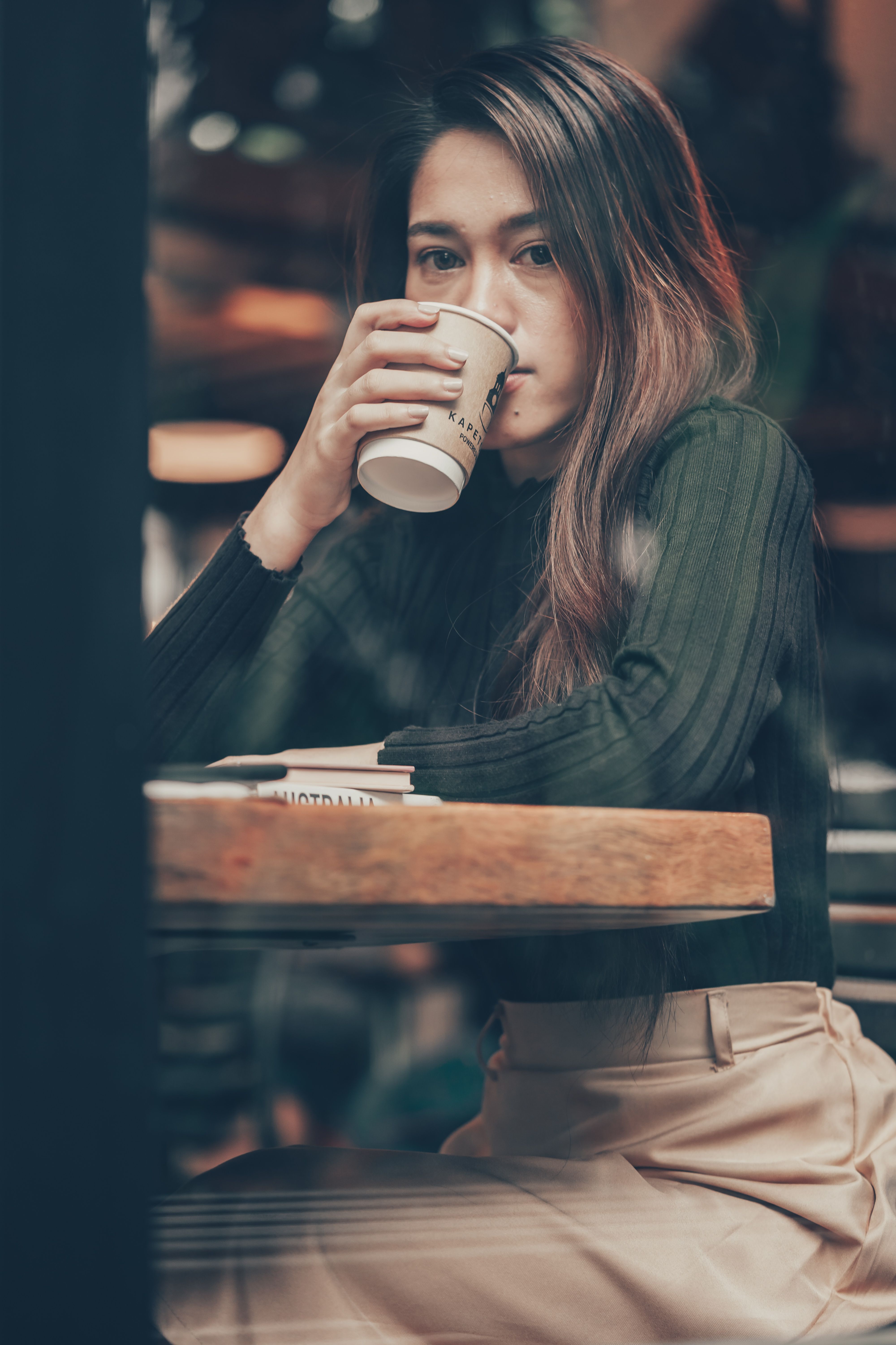 Asian woman enjoying a hot beverage in a cozy cafe, captured through a glass for a candid vibe.