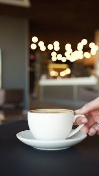 Hand holding coffee cup with bokeh lights in cozy cafe setting, perfect for lifestyle themes.