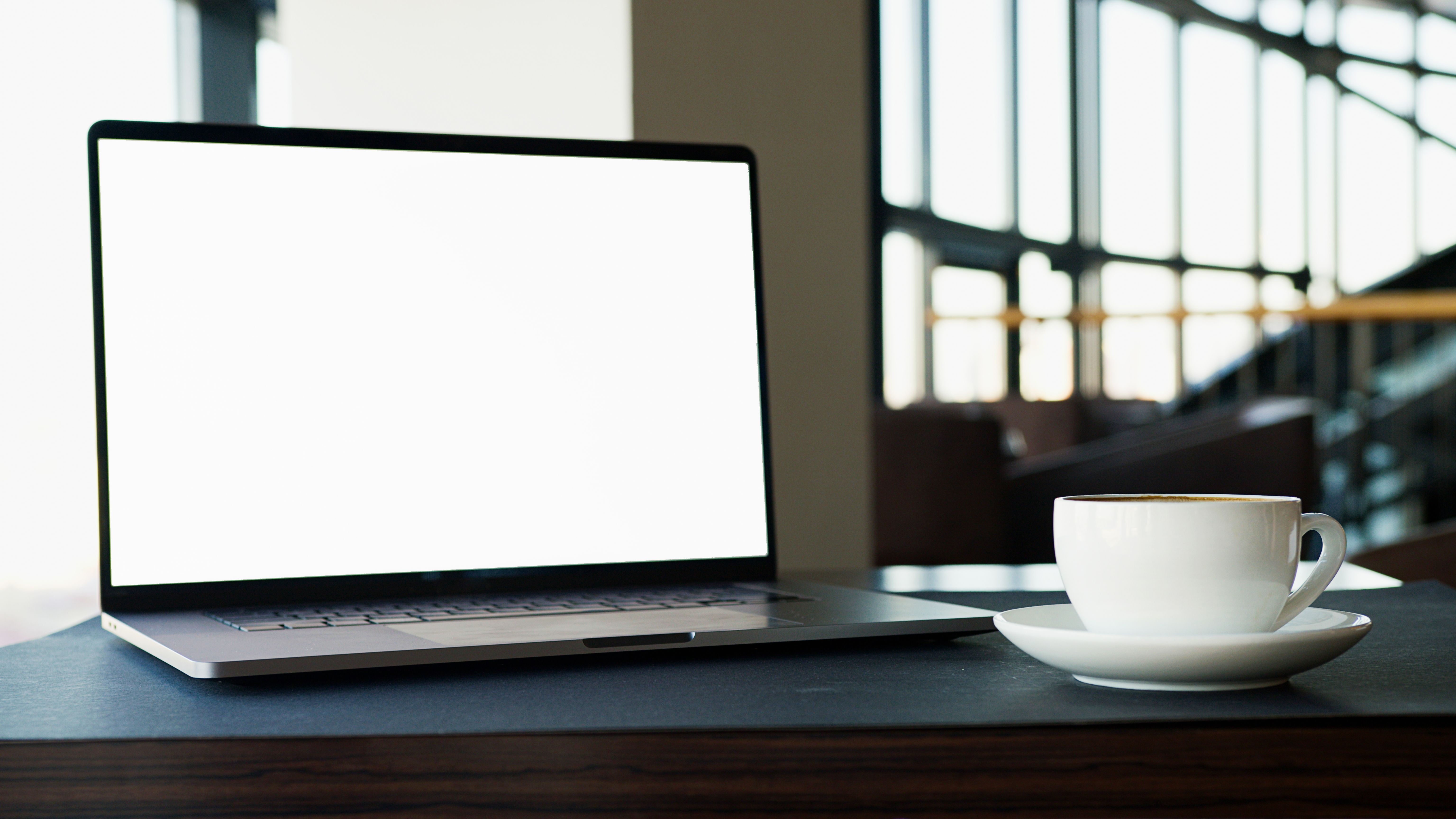 A blank-screened laptop beside a steaming coffee cup on a desk in a modern office setting.