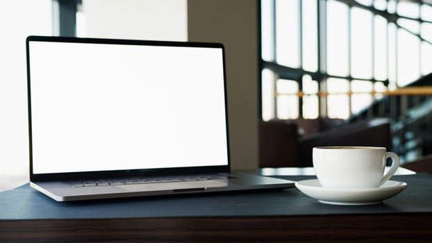 A blank-screened laptop beside a steaming coffee cup on a desk in a modern office setting.