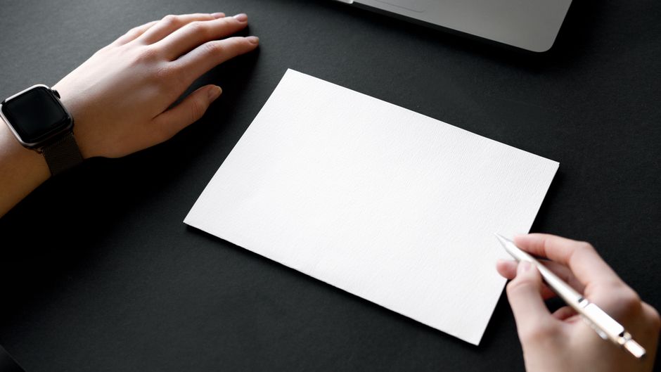 Hands positioned over blank paper ready to write with a pen, accompanied by a laptop.