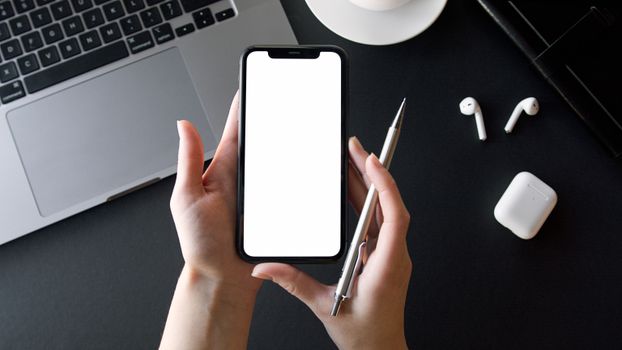Close up of hands holding a smartphone over a desk with laptop and earbuds.