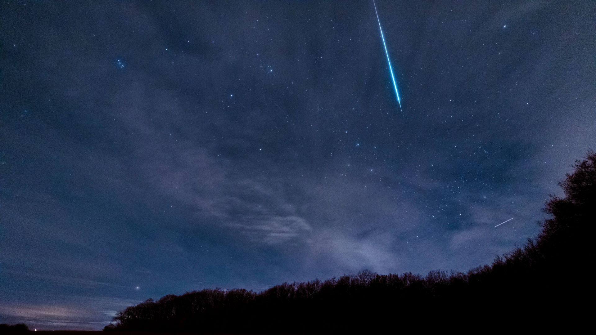 Breathtaking view of a meteor streaking across a starry and cloudy night sky above dark silhouettes of trees.