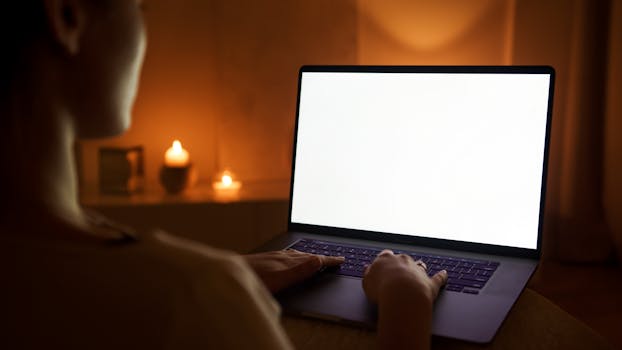 A person working on a laptop with a blank screen, illuminated by candlelight.
