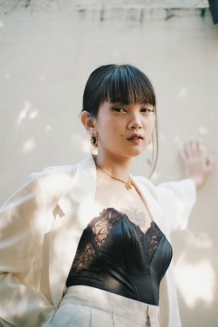 Trendy young Asian female in black corset and white jacket standing in street near white wall and looking at camera in daytime