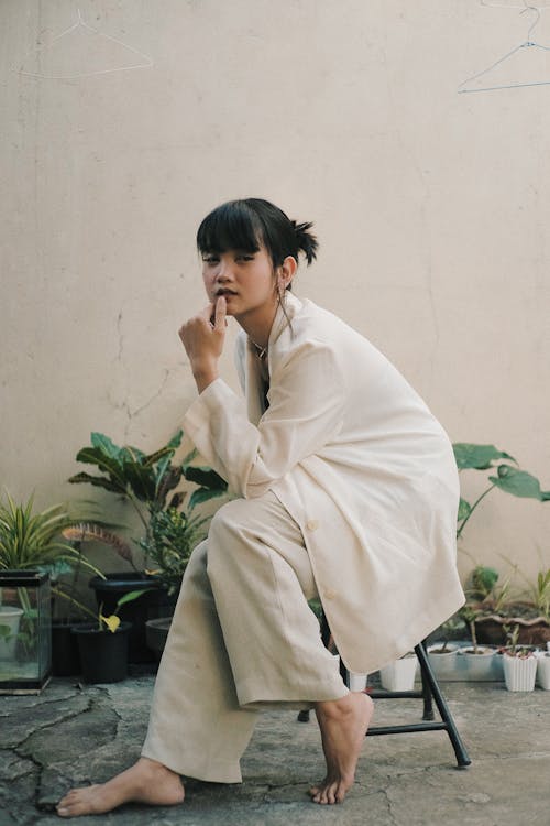 Full body of confident barefoot young ethnic woman in stylish white outfit sitting on chair in street near white wall and plants in pots and looking at camera in daylight