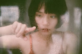 Through window of young Asian woman drawing on  wet glass with finger in soft light