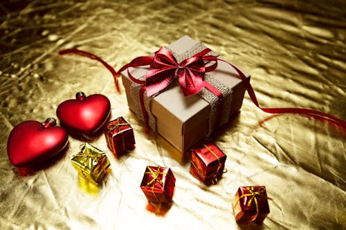 From above of various wrapped gift boxes with bows placed on shiny golden surface with red heart shaped Christmas tree baubles