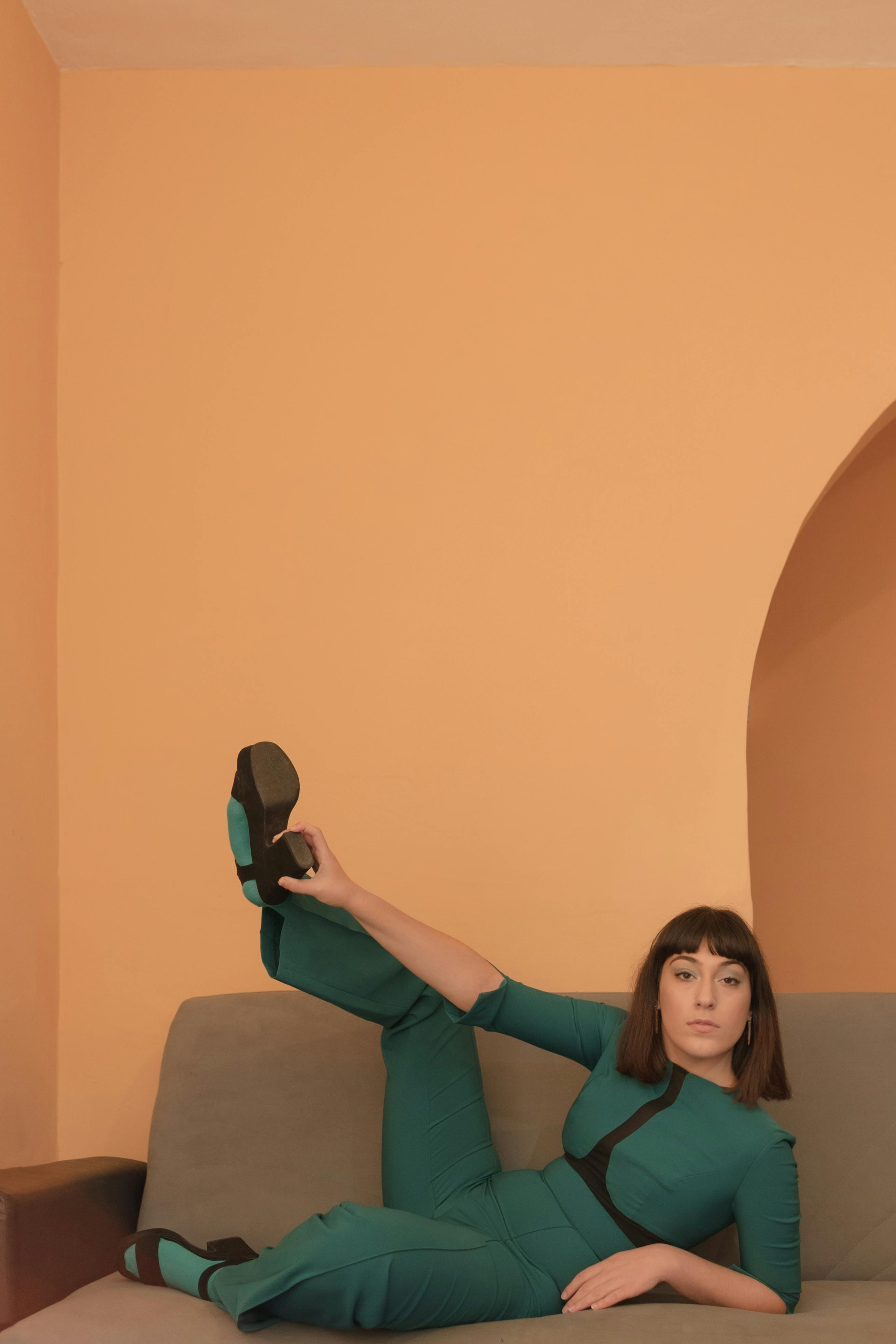 Woman in green outfit striking a pose on a couch against an orange wall.