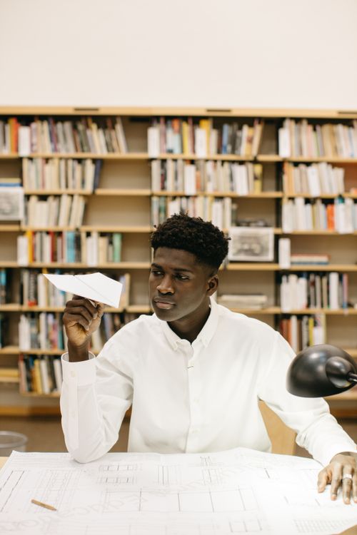 Free A thoughtful student holds a paper plane in a library filled with books, showcasing creativity. Stock Photo