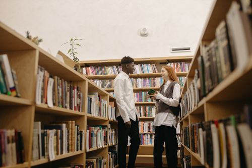 Free Two students exchange smiles and ideas between library bookshelves. Stock Photo