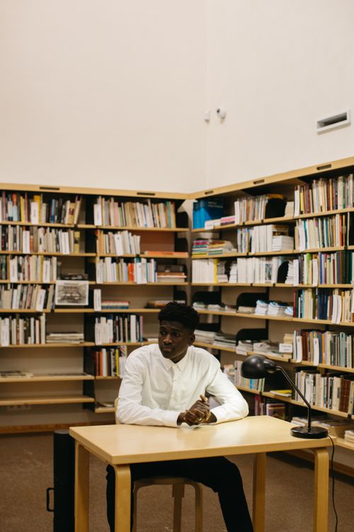 Free A young man in a library sitting at a desk, surrounded by books, deep in thought. Stock Photo