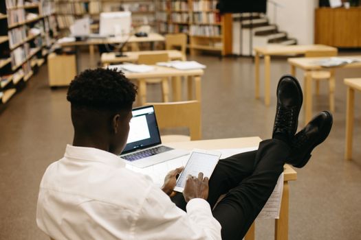Black man studying in library with laptop and tablet, feet up on desk.