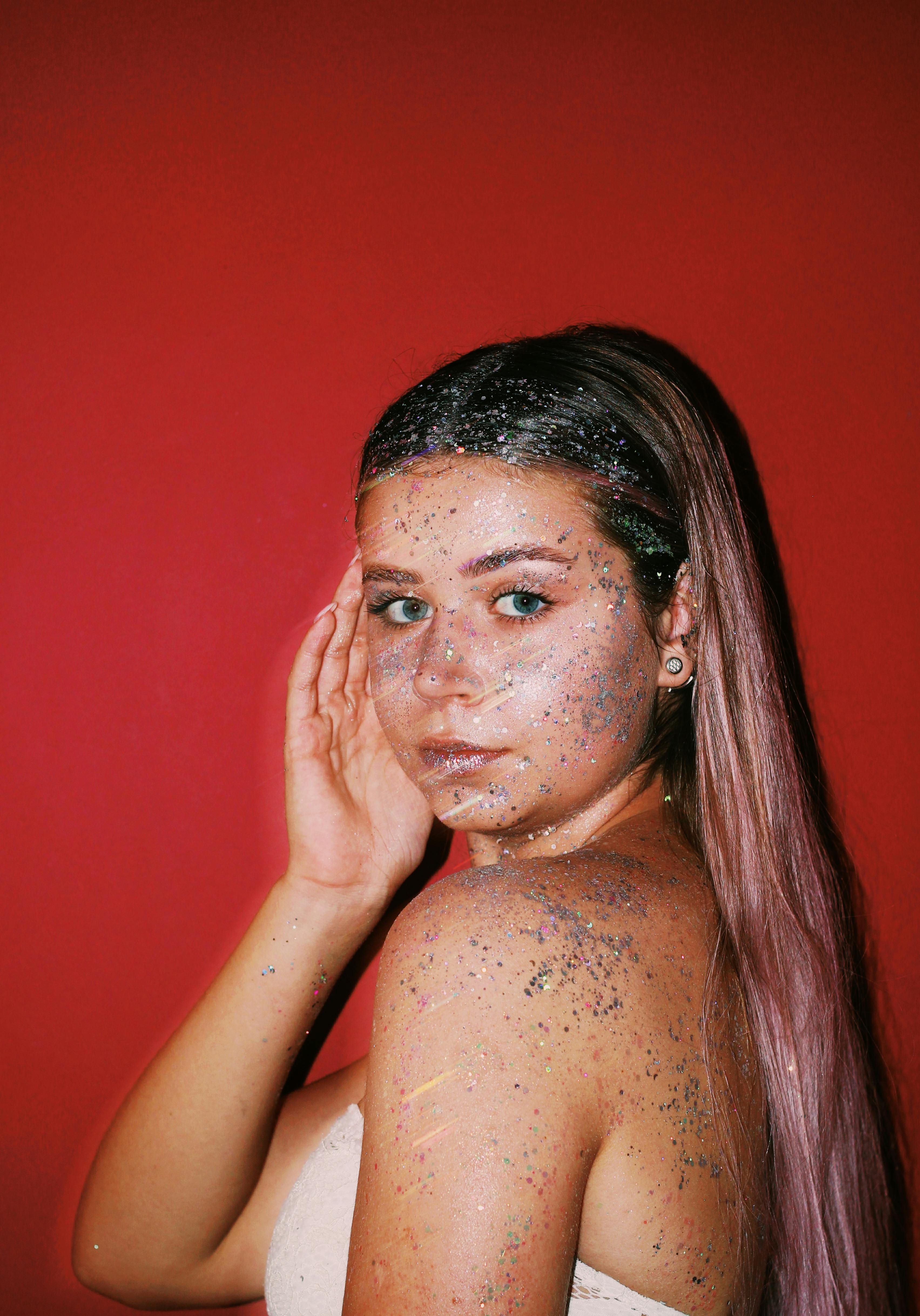 A woman with glitter on her skin poses against a vivid red backdrop.