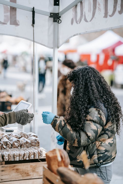 Side view of unrecognizable curly female customer in jacket and protective gloves paying for purchase by card in street food market