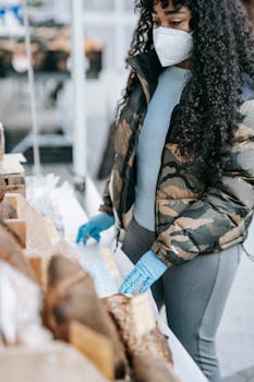 Woman wearing mask and gloves selecting bread at outdoor market stall during daytime.