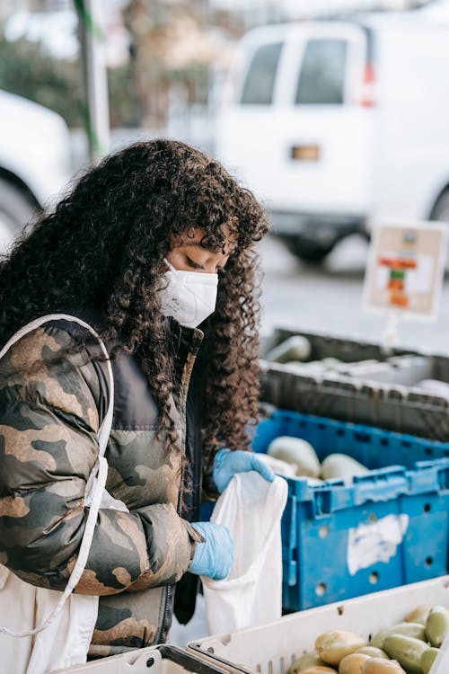 Side view of young  African American female with in protective mask and rubber gloves putting fruits in bag in outdoor grocery store