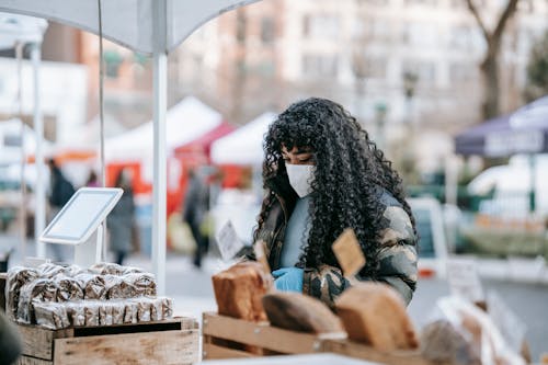 Young African American female in warm casual outfit and medical mask looking at showcase with various breads at street fair