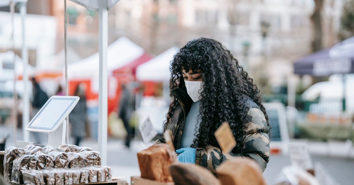 Young African American female in warm casual outfit and medical mask looking at showcase with various breads at street fair