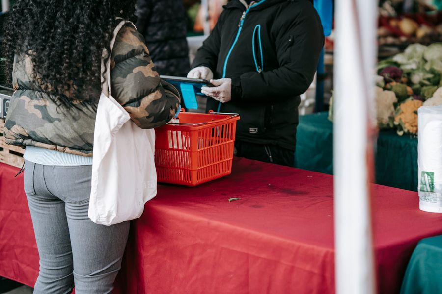 Crop anonymous seller in protective gloves calculating cost while assisting female customer at counter in local farm market during coronavirus pandemic