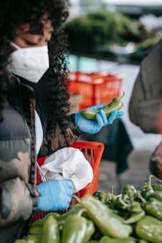 Crop concentrated African American female in respirator and gloves picking ripe capsicums while doing groceries in supermarket