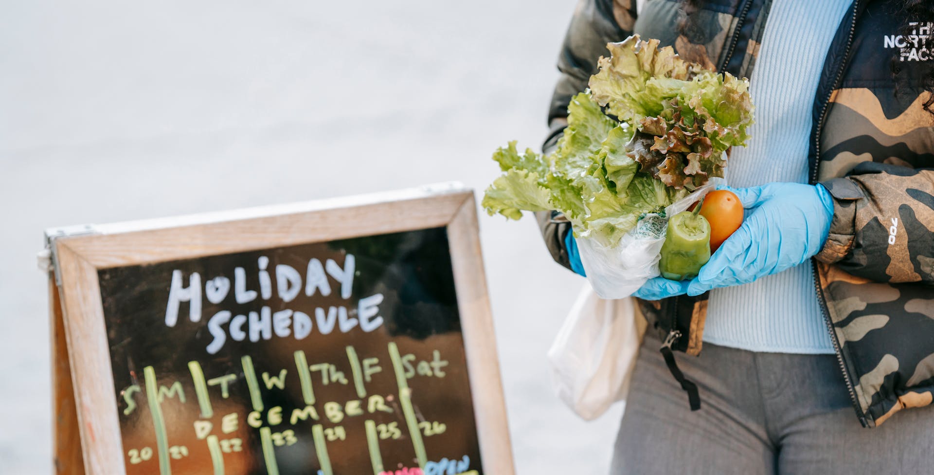 Crop anonymous female in warm jacket and protective gloves showing fresh ripe vegetables while standing near grocery chalkboard on street