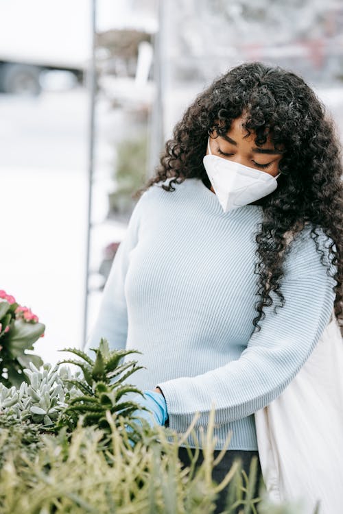 Focused African American female in respirator and gloves picking verdant potted plants in outdoor floral market